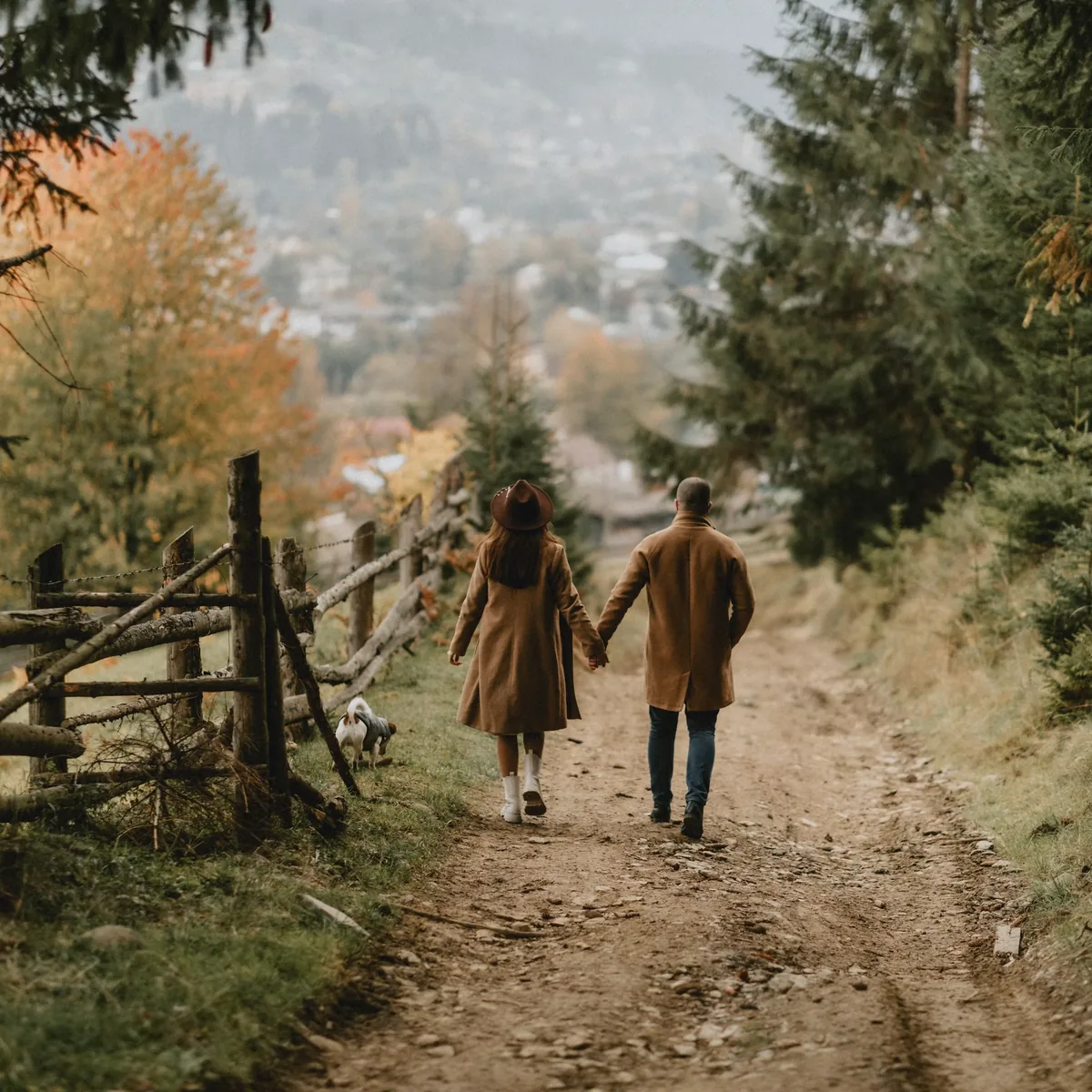 A couple walking a forest path with their dog toward autumn hills
